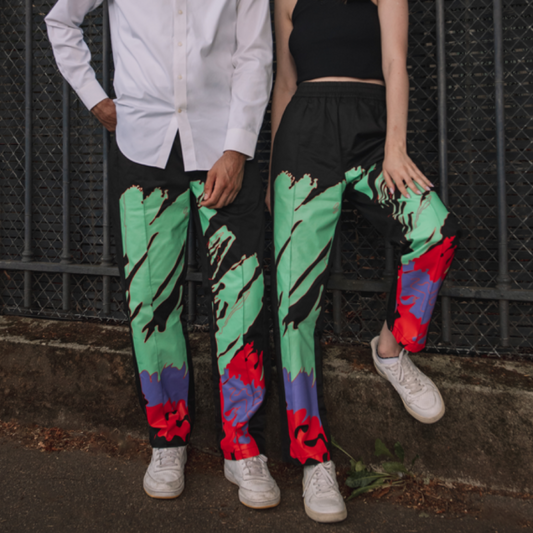 Two individuals standing side by side against a fence, wearing colorful patterned trousers and white tops.