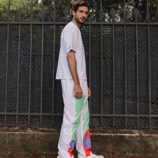A person in a white outfit with colorful patterns on the trousers standing in front of a fence.